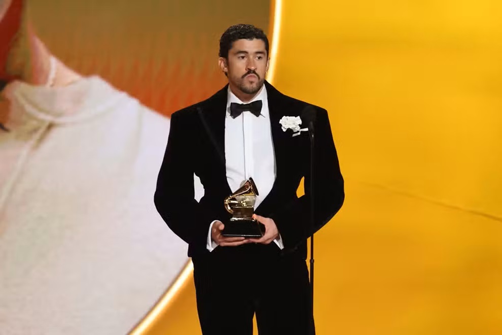 Quem é Bad Bunny? Artista ganhou Grammy 2026 6 Bad Bunny de terno segurando troféu Grammy em palco dourado, expressão de conquista
