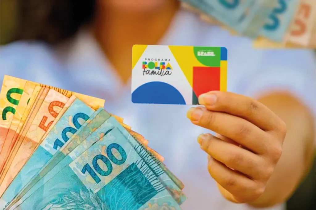 Bolsa Família de Fevereiro continua com os pagamentos nesta semana; veja quem recebe 6 Beneficiária segura cartão do Bolsa Família com notas de reais ilustrando o calendário de pagamentos entre 23 e 27 de fevereiro.