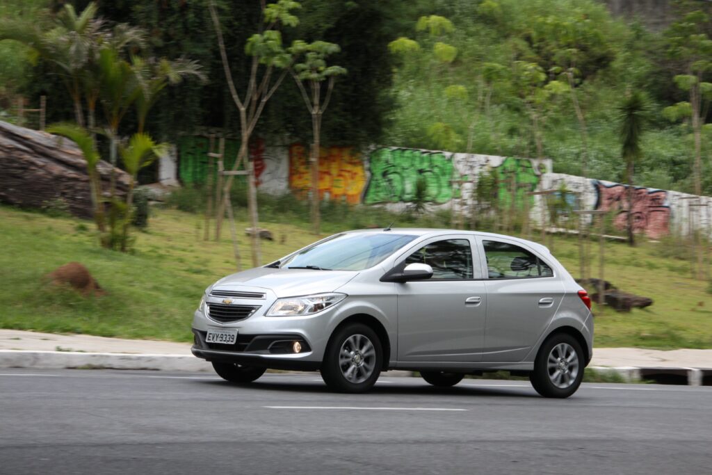 Os 5 modelos de carros usados que menos perderam valor em 2025 7 Carro hatch prata circula por rua asfaltada com muro grafitado e vegetação lateral