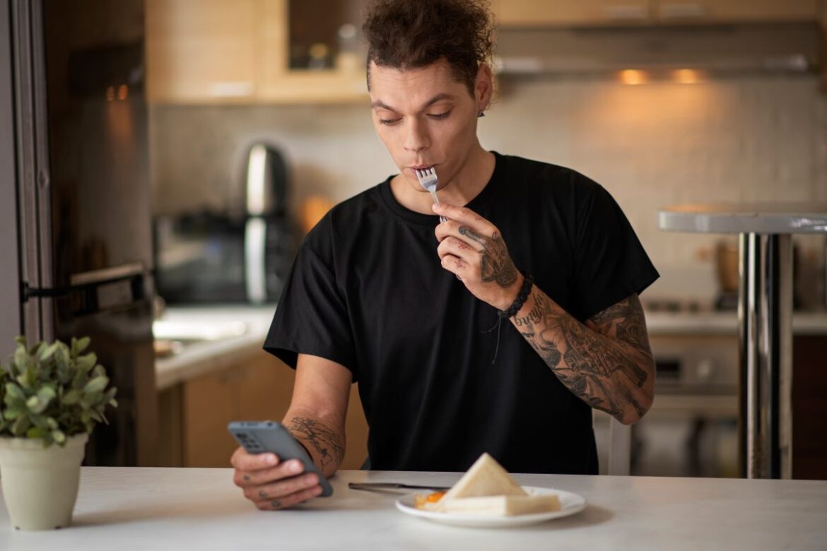 Homem jovem com tatuagens no braço sentado à mesa de uma cozinha iluminada, segurando um garfo na boca enquanto olha atentamente para a tela de um celular na outra mão. À sua frente, um prato com sanduíche.
