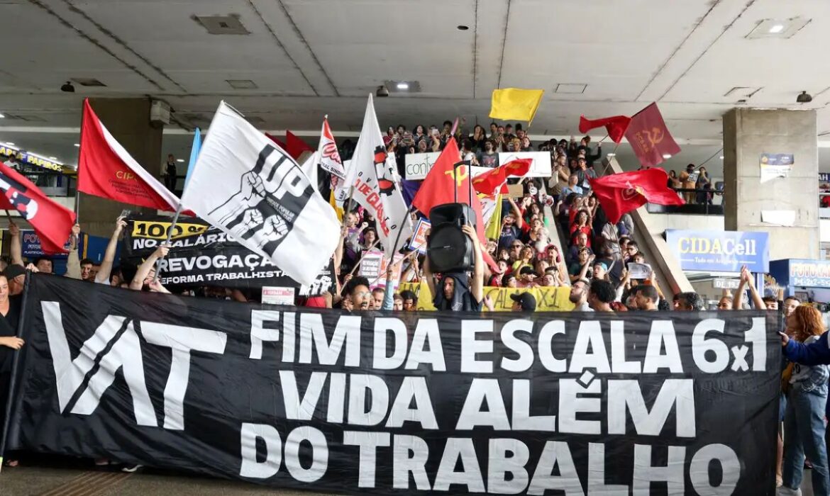 Manifestação popular em um local coberto com escadarias ao fundo. No primeiro plano, uma grande faixa preta com letras brancas diz: "VAT - FIM DA ESCALA 6x1 - VIDA ALÉM DO TRABALHO". Diversas pessoas seguram bandeiras vermelhas e brancas de movimentos sociais e sindicais enquanto protestam.