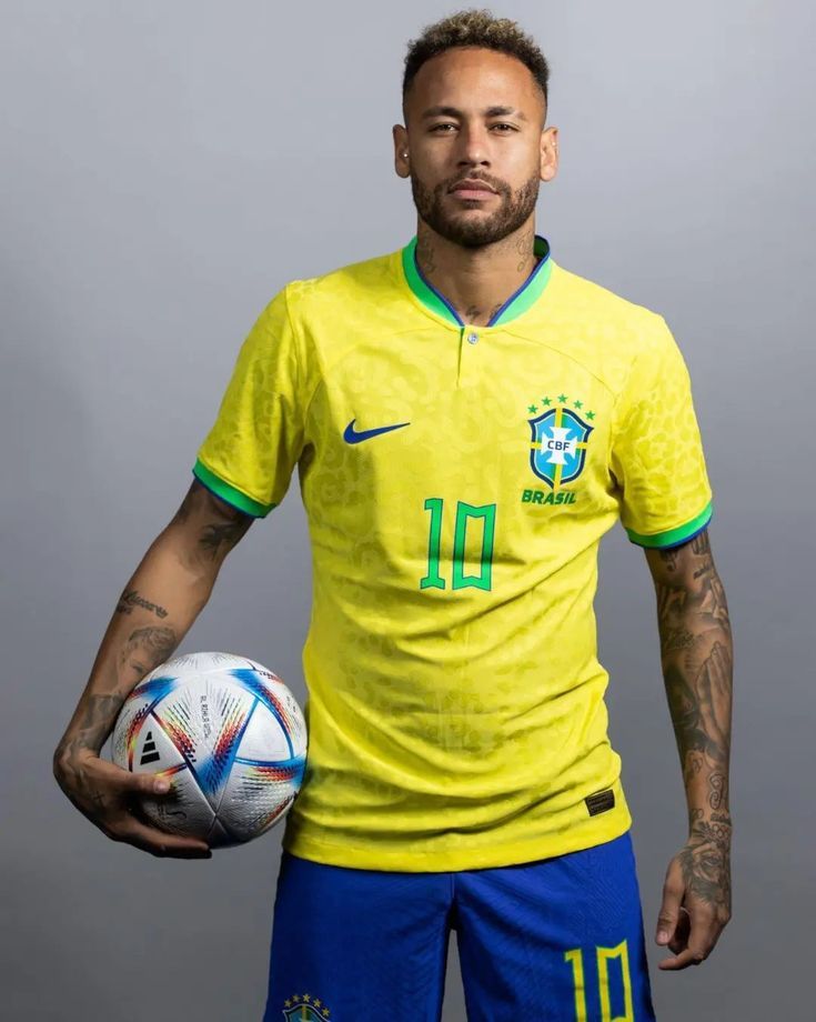 Neymar faz 34 anos hoje: recuperação de lesão e sonho da Copa marcam momento 6 Neymar Jr. posa de pé, segurando uma bola de futebol oficial da Copa do Mundo sob o braço direito. Ele veste o uniforme principal da Seleção Brasileira (amarelo com detalhes verdes), exibindo o número 10 verde no peito e diversas tatuagens nos dois braços. O fundo é de cor cinza neutro.