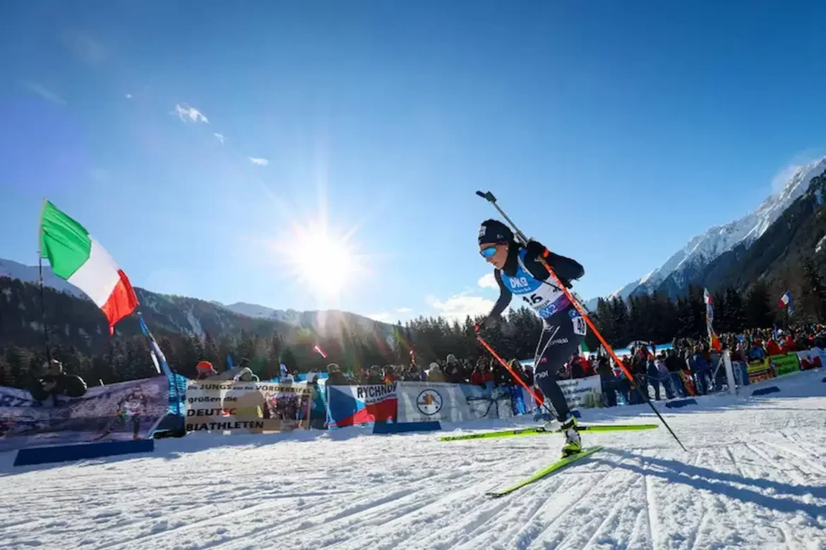 Rebecca Passler competindo no esqui de fundo durante uma prova no inverno, com bandeiras de vários países ao fundo, em uma paisagem montanhosa.