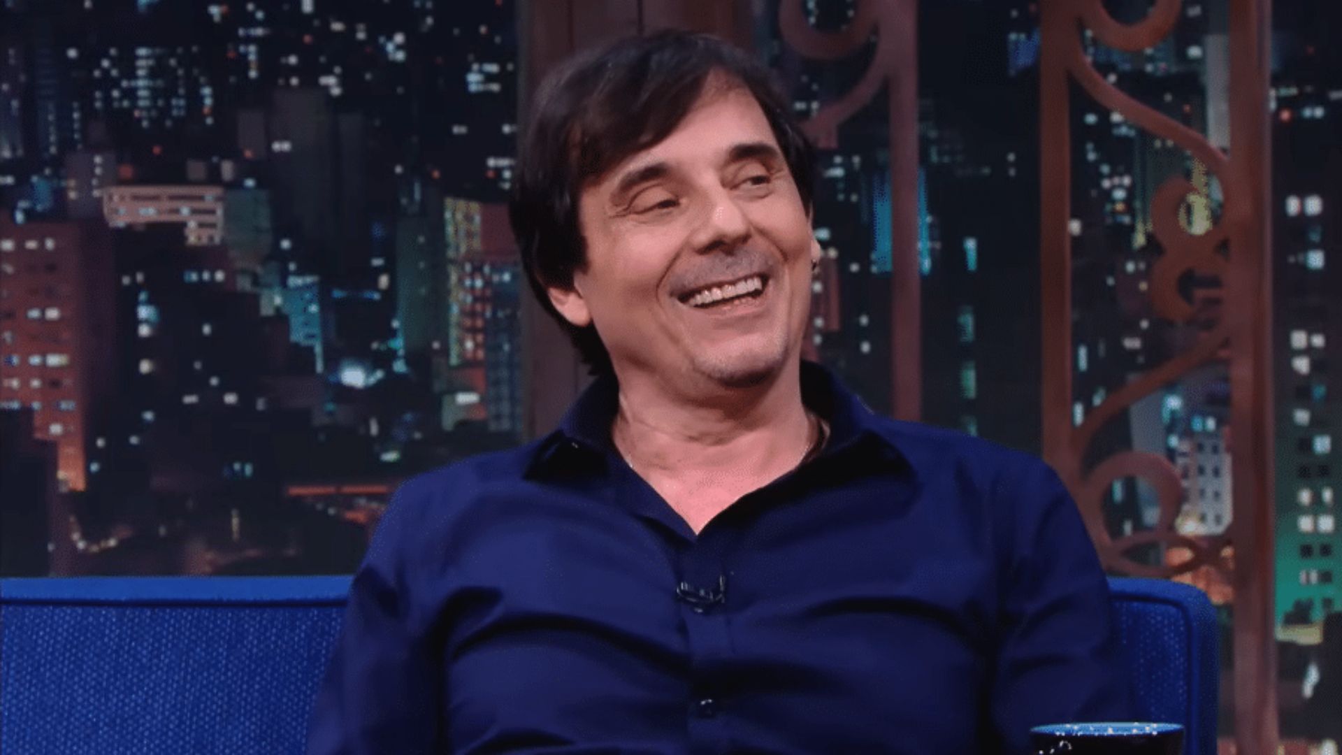 Luto no mundo da dublagem: Ricardo Schnetzer, voz de Tom Cruise e Al Pacino, morre aos 72 anos 6 Ricardo Schnetzer sorrindo, sentado em um sofá azul durante uma entrevista em estúdio de televisão. Ele veste uma camisa social azul escura, tem cabelos castanhos e o fundo mostra luzes de uma cidade à noite através de janelas decorativas.