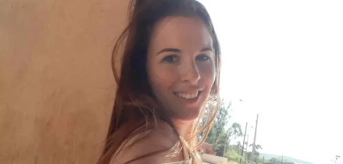 Mulher de cabelos castanhos sorri ao ar livre próxima a parede em ambiente rural, céu claro ao fundo