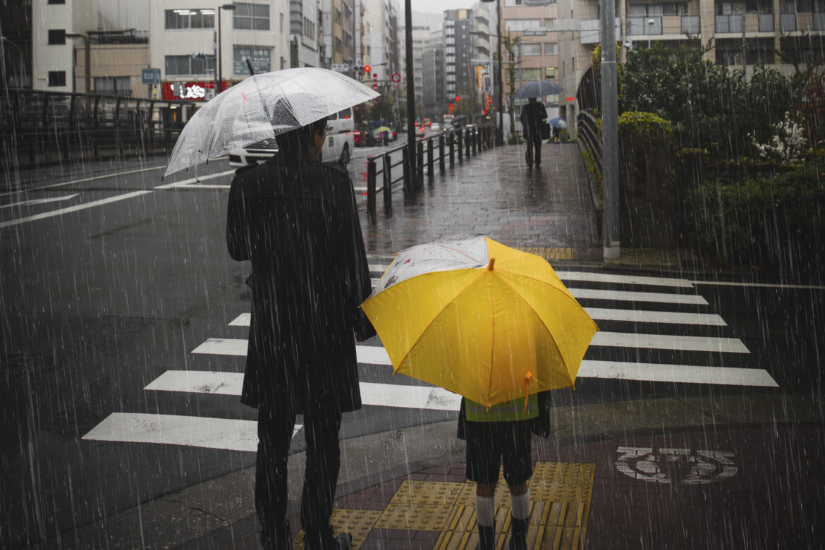 Família caminhando sob forte chuva em uma rua, protegidos por guarda-chuvas em um cenário urbano.