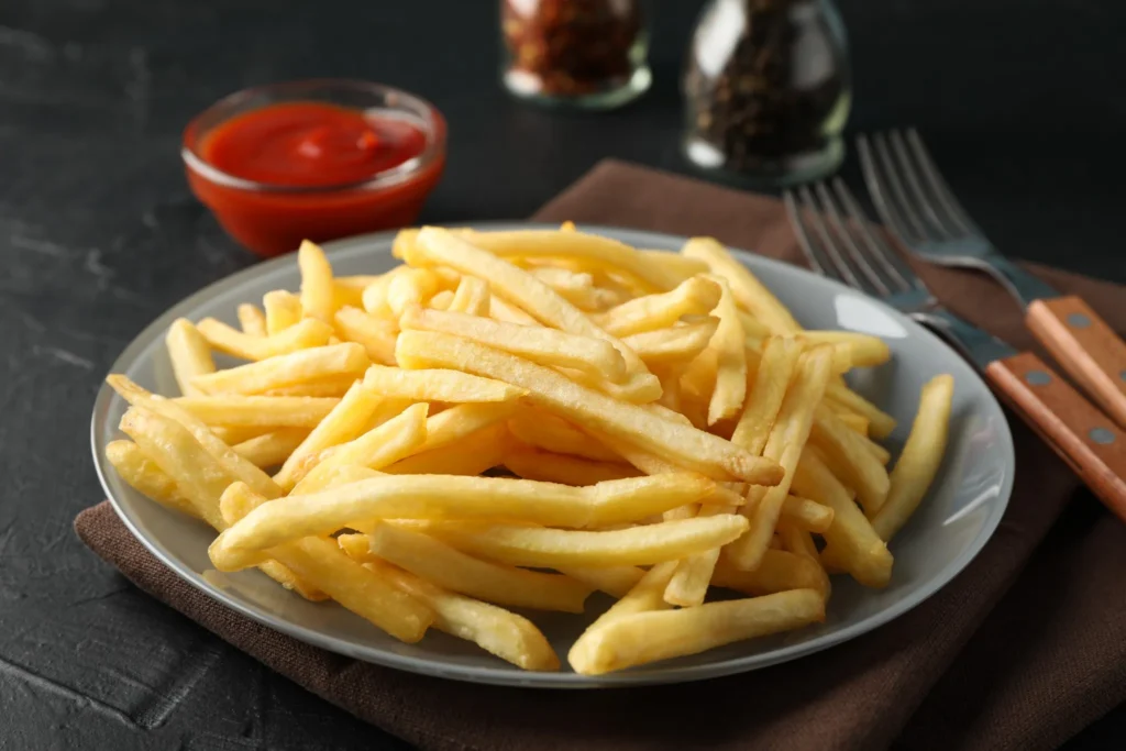 Batata frita de cinema: O ingrediente seco que você polvilha antes de fritar para deixá-la crocante por horas 6 Imagem de batatas fritas caseiras crocantes em um prato, com molho de ketchup ao fundo e utensílios de mesa.