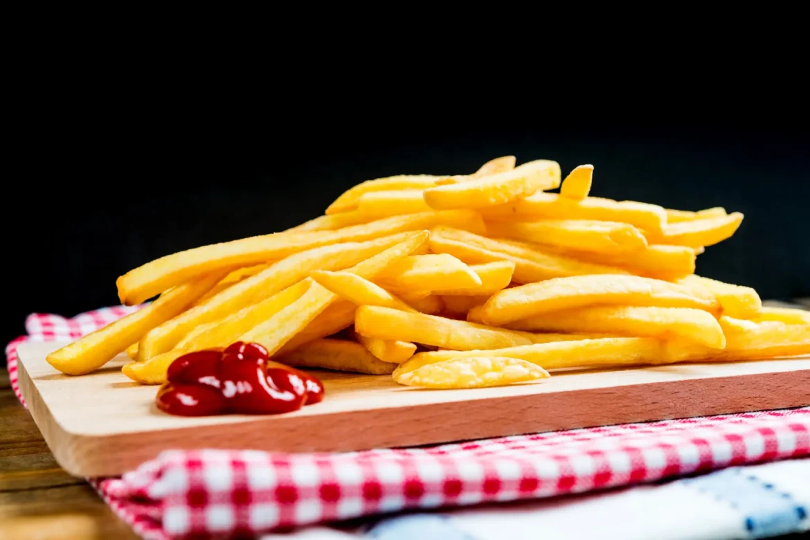 Imagem de batatas fritas crocantes em uma tábua de madeira com molho de ketchup, com fundo escuro.