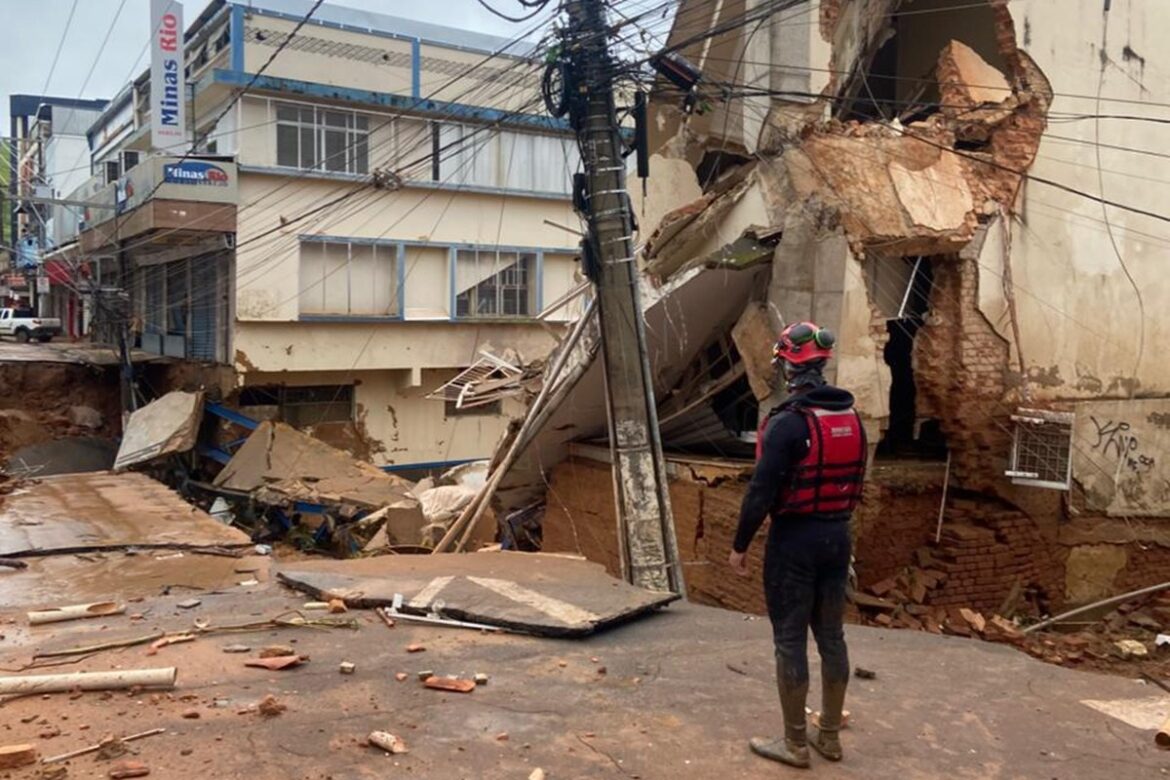 Agente da defesa civil observa edifício destruído após desabamento em rua alagada