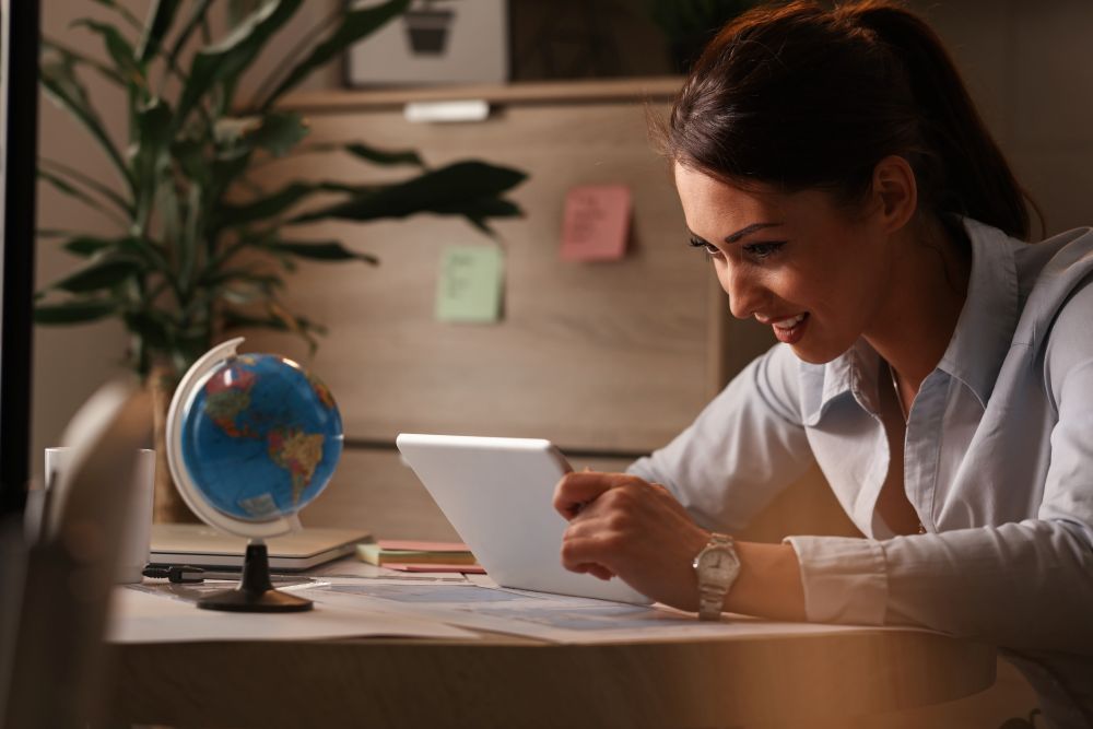 A terça-feira mágica: Por que você deve procurar passagens aéreas às 15h 6 Mulher jovem e sorridente, sentada à mesa em um ambiente aconchegante à noite, utilizando um tablet branco para pesquisar voos. À sua frente, há um pequeno globo terrestre azul e documentos de viagem, iluminados por uma luz quente de luminária, simbolizando o planejamento de férias.