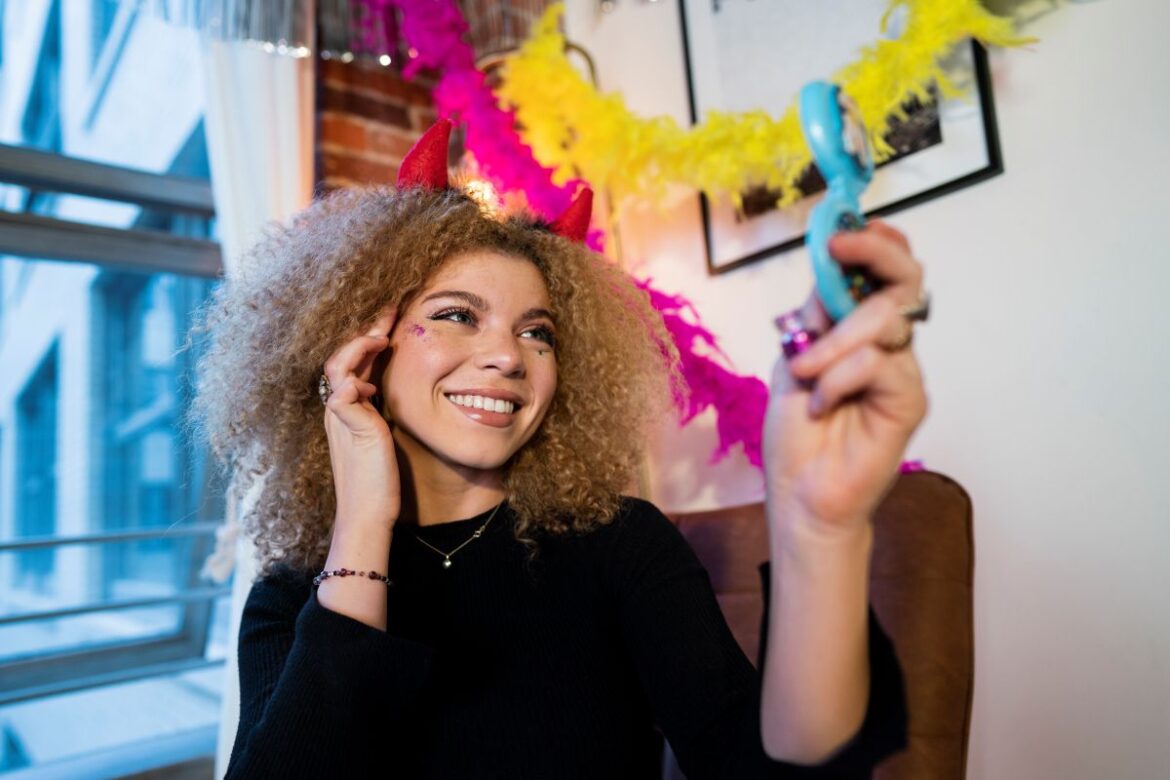 Uma mulher jovem sorridente com cabelo cacheado volumoso e tiara de chifres vermelhos. Ela usa brilho no rosto e olha para um espelho azul enquanto se prepara para o Carnaval. Ao fundo, há enfeites de plumas coloridas em rosa e amarelo.