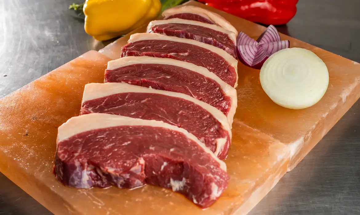 Carne de segunda com gosto de filé: O ingrediente de 3 reais que amacia qualquer corte em apenas 20 minutos 6 Fatias de carne bovina crua alinhadas em tábua de sal com cebola e pimentão ao fundo