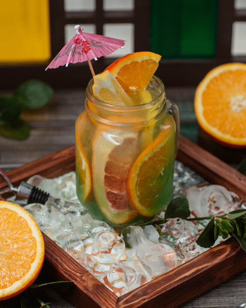 Drinks refrescantes: 5 receitas de coquetéis sem álcool para o verão 2026 8 Um pote de vidro estilo "mason jar" preenchido com chá gelado translúcido, contendo fatias generosas de laranja e toranja mergulhadas entre cubos de gelo. O drink é decorado com um pequeno guarda-sol rosa no topo e está sobre uma bandeja de madeira rústica cercada por mais gelo e laranjas cortadas.