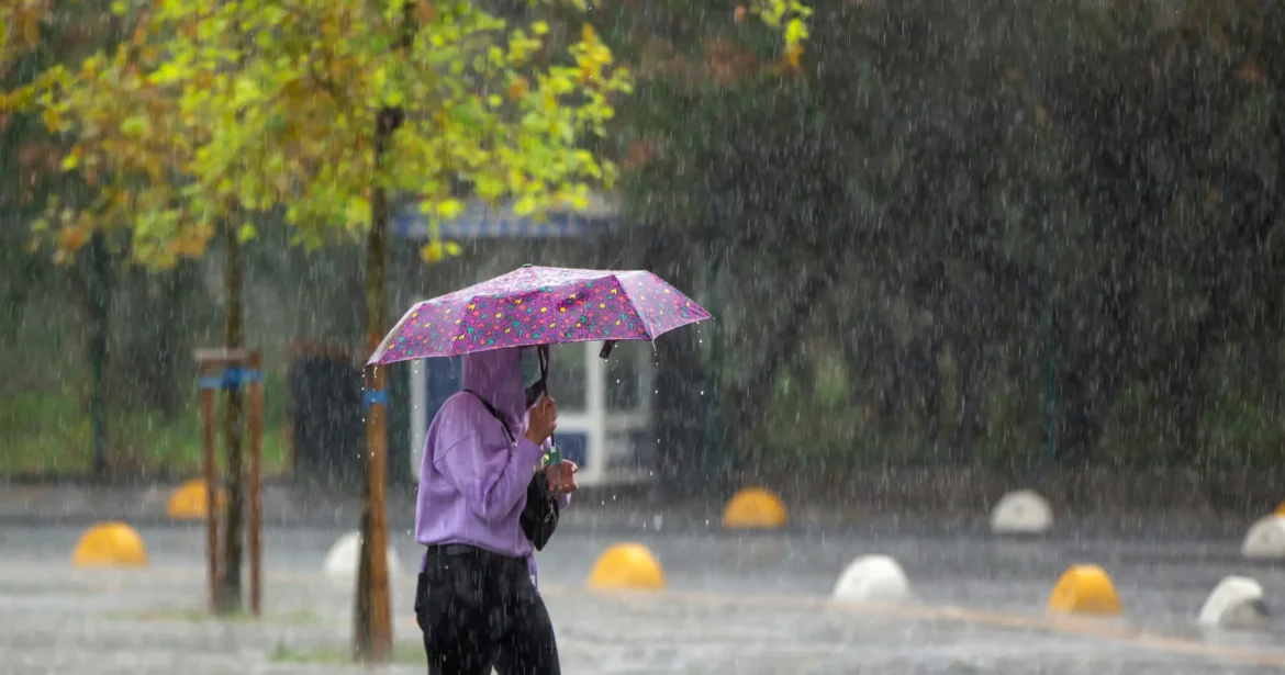 Pessoa com guarda-chuva colorido caminhando sob chuva intensa em área urbana.