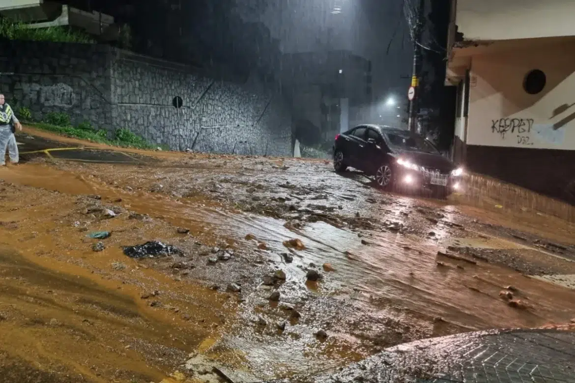 Foto noturna de uma rua íngreme em Juiz de Fora sob chuva forte, com enxurrada de lama e pedras descendo a via e um carro preto com faróis acesos parado no meio da inundação.