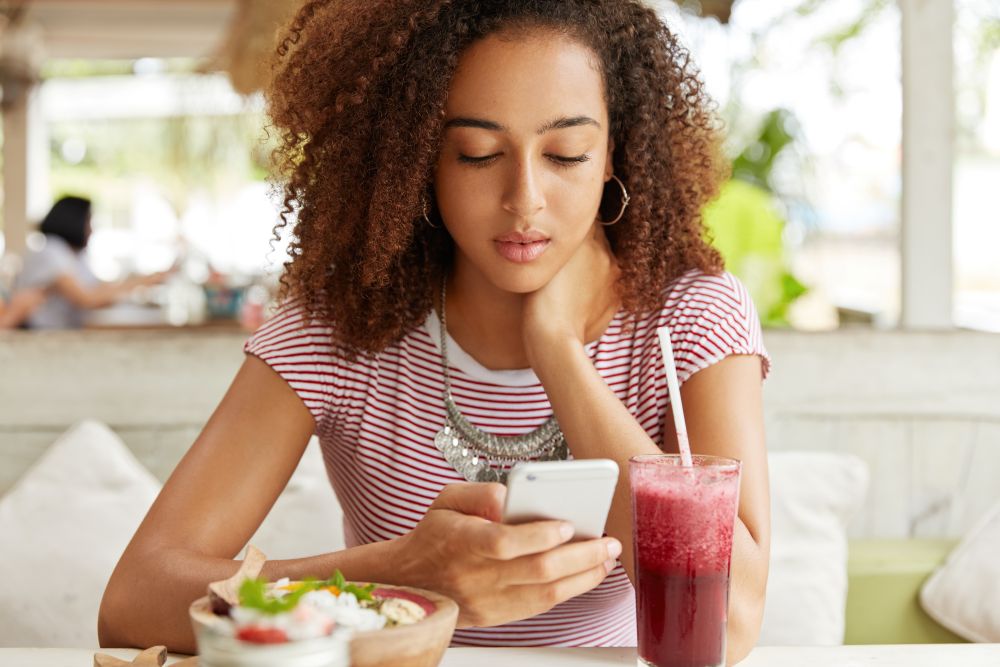 Por que você come sem fome enquanto olha o celular? Veja como quebrar esse ciclo hoje 6 Jovem mulher de cabelos cacheados, usando argolas e colar, sentada à mesa de um restaurante enquanto digita no celular. À sua frente, há uma tigela de açaí com frutas e um copo de suco vermelho com canudo.