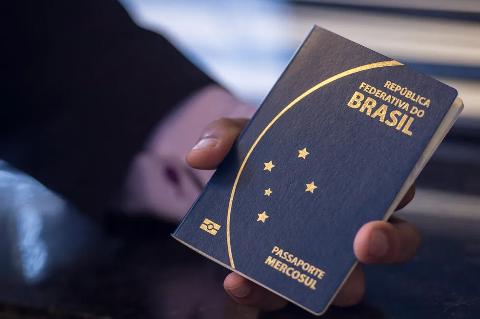 Como obter o passaporte: confira passo a passo 6 Pessoa segura passaporte brasileiro sobre balcão escuro durante solicitação do documento
