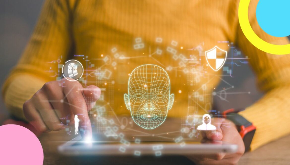 Close-up de mãos em um suéter amarelo utilizando um tablet, onde uma sobreposição digital exibe um modelo em grade 3D de um rosto humano, ícones de cadeado e impressão digital, em meio a bolhas coloridas rosa, amarela e azul.