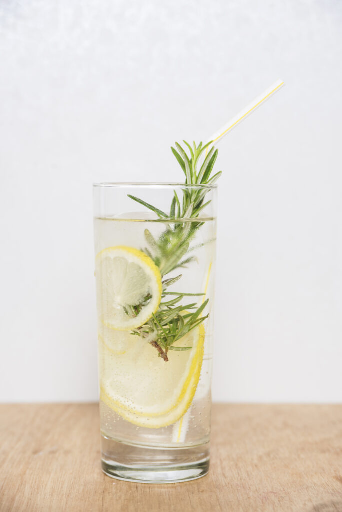Drinks refrescantes: 5 receitas de coquetéis sem álcool para o verão 2026 7 Um copo alto de vidro transparente contendo um drink translúcido e efervescente com rodelas de limão siciliano e um ramo generoso de alecrim fresco. O copo tem um canudo listrado de amarelo e branco, gotas de condensação na parte externa e está posicionado sobre uma superfície de madeira clara contra um fundo branco.