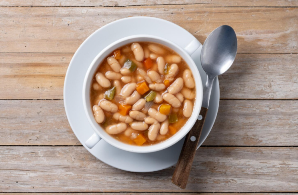 Vista superior de uma tigela de cerâmica branca com duas alças contendo uma sopa de feijão branco cozido. O caldo é encorpado e inclui cubos de cenoura alaranjada e pedaços de pimentão verde. Uma colher de metal com cabo de madeira repousa ao lado do prato sobre uma mesa de madeira rústica clara.