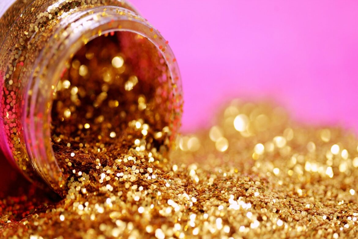 Pote de glitter dourado derramado sobre superfície rosa, partículas brilhando