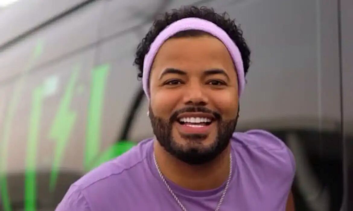 Influenciador Hytalo Santos, sorrindo e usando uma faixa lilás na cabeça, em frente a um veículo escuro com detalhes em verde.