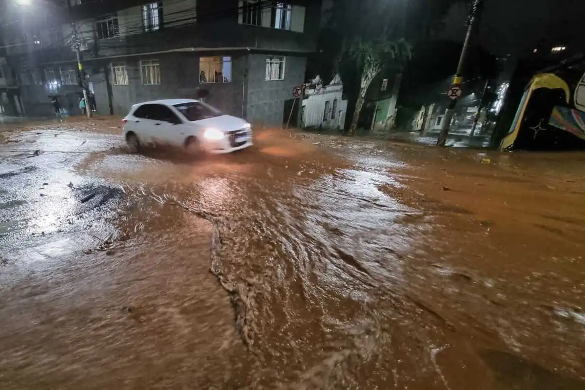Chuva intensa causa destruição em Juiz de Fora (MG); veja imagens impressionantes 7 Rua inundada por lama em Juiz de Fora durante a noite; à esquerda, uma pessoa sob um guarda-chuva observa a via obstruída por galhos e troncos, com dois carros brancos parados em meio aos destroços.