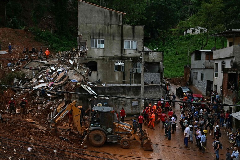 Vista de um cenário de devastação após deslizamento de terra em Minas Gerais, mostrando uma casa parcialmente destruída e equipes de resgate operando uma retroescavadeira em meio à lama e escombros.