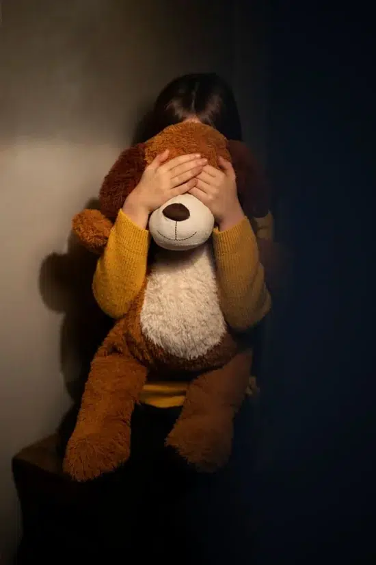 Criança com suéter amarelo cobre o rosto de um urso de pelúcia em ambiente escuro, simbolizando vítimas de investigação sobre rede de abuso sexual infantil ligada a piloto preso.