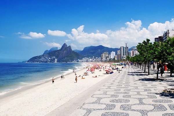 Carnaval 2026: guia completo dos melhores lugares para curtir a folia no Brasil 6 Vista da Praia de Ipanema em um dia ensolarado, mostrando o calçadão de pedras portuguesas com padrão de ondas em primeiro plano. A areia branca está repleta de guarda-sóis coloridos e banhistas, com o mar azul à esquerda e o Morro Dois Irmãos ao fundo sob um céu limpo.