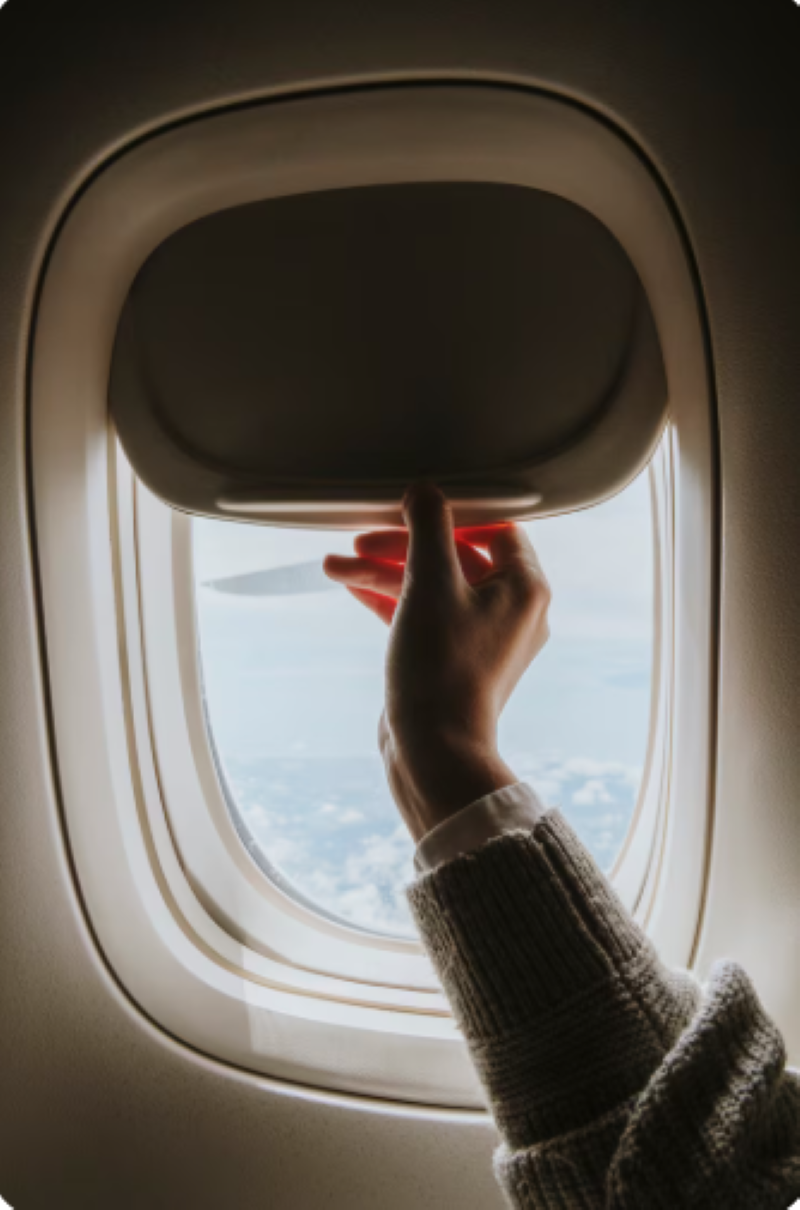 Por que as janelas dos aviões são redondas? O motivo de engenharia que salva vidas diariamente 6 Close-up vertical do interior de uma cabine de avião. Uma mão feminina, vestindo um casaco de lã cinza, puxa a persiana bege de uma janela arredondada. Pela abertura, vê-se a asa da aeronave sobre um mar de nuvens brancas sob um céu claro.