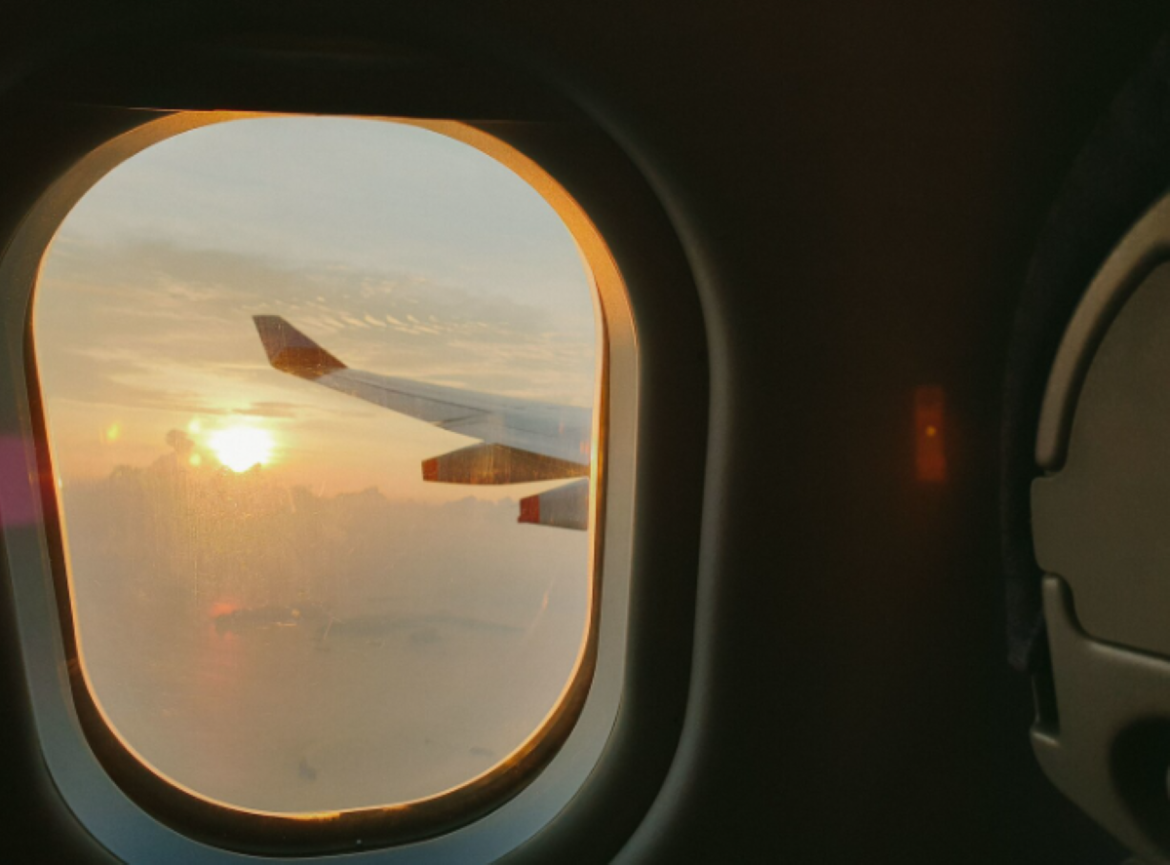 Vista interna de uma janela de avião com bordas arredondadas. Através do vidro, observa-se a asa da aeronave sobre nuvens durante o pôr do sol, com tons alaranjados refletindo na estrutura metálica. À direita, parte de uma poltrona de passageiro é visível no interior escurecido da cabine.