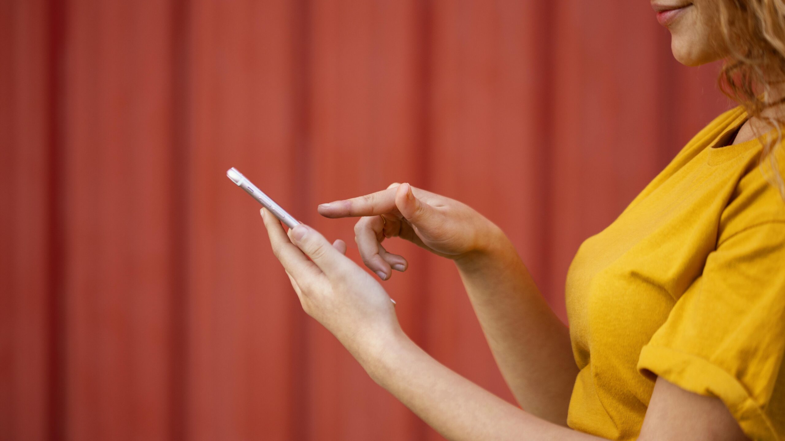 Apple prepara iPhone 17e, sua versão mais acessível; veja o que esperar 6 Mulher com camiseta amarela usando smartphone próximo a parede vermelha