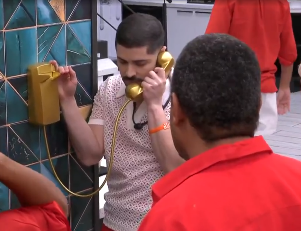 BBB 26: veja quem atendeu cada uma das seis ligações do Big Fone no primeiro mês 6 O participante Marcelo, vestindo uma camisa polo branca de renda, atende ao Big Fone dourado preso em uma parede de azulejos geométricos azuis, enquanto outros participantes de vermelho observam.