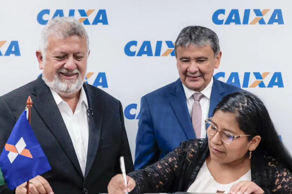 Mulher assinando documento enquanto dois homens observam sorrindo diante de painel da Caixa