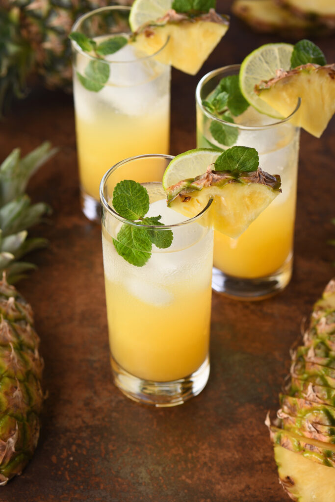 Drinks refrescantes: 5 receitas de coquetéis sem álcool para o verão 2026 6 Três copos longos de vidro servidos com mojito de abacaxi amarelo vibrante e gelo. Cada drink está decorado com uma rodela de limão, um triângulo de abacaxi fresco na borda e ramos de hortelã verde. Ao lado dos copos, há pedaços de abacaxi com casca sobre uma superfície rústica.