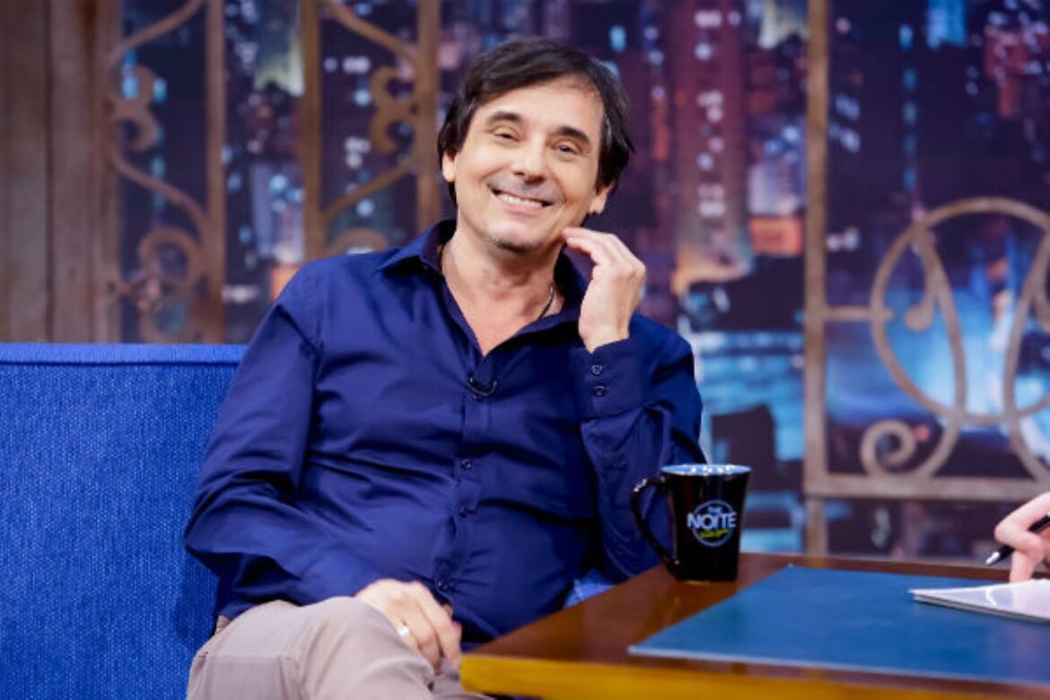 Ricardo Schnetzer sorri enquanto está sentado em um sofá azul durante uma entrevista em um cenário de talk show noturno. Ele veste uma camisa azul escura de mangas compridas, tem cabelos escuros e a mão esquerda encostada no queixo. À sua frente, há uma caneca preta com o logotipo do programa sobre uma mesa de madeira.
