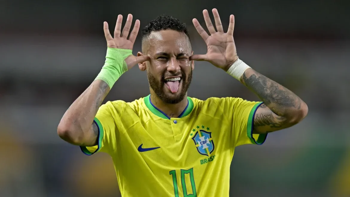 Neymar Jr. com a camisa amarela da Seleção Brasileira fazendo sua comemoração característica: língua para fora e mãos abertas ao lado do rosto. Ele possui uma bandagem verde no punho direito, tatuagens nos braços e um sorriso vibrante durante uma partida oficial.