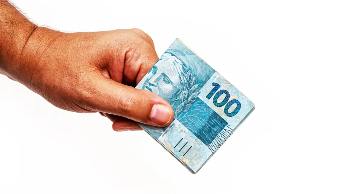 Homem segurando nota de R$ 100 sobre fundo branco.