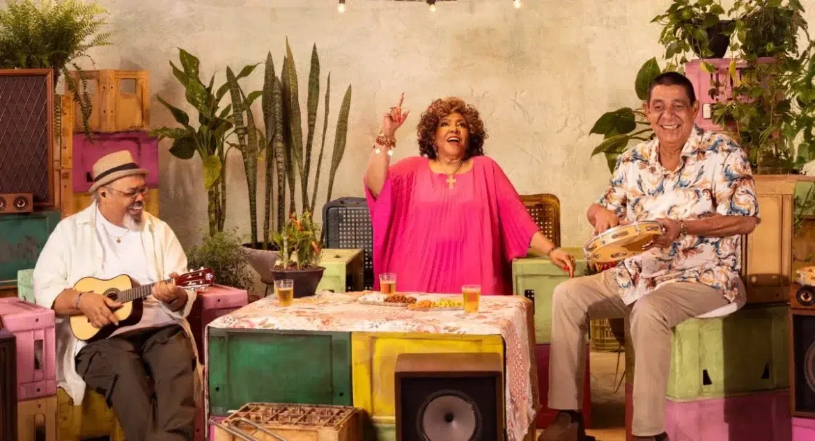 Três pessoas sorriem em roda de samba, com instrumentos e bebidas sobre mesa colorida