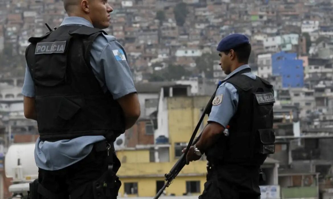 Dois policiais militares fardados, de costas, observando uma área urbana densamente povoada em uma encosta de morro.