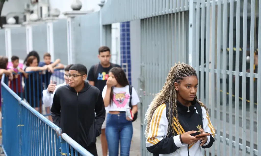 Prouni 2026: veja se você está entre os 226 mil pré-selecionados na primeira chamada 6 Estudantes em fila próximos a grade de escola, segurando documentos e celulares