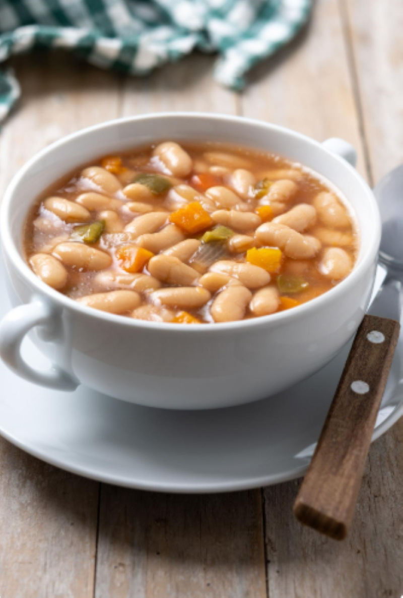 O segredo do feijão: O que colocar na água do molho para nunca mais sentir gases ou estufamento 6 Close-up de uma sopa de feijão branco servida em uma tigela de cerâmica branca sobre uma mesa de madeira. O prato contém grãos inteiros, cubos de cenoura e pimentão verde em um caldo suculento. Ao fundo, um pano de prato xadrez verde e branco aparece levemente desfocado.