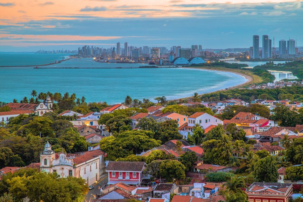 Carnaval 2026: guia completo dos melhores lugares para curtir a folia no Brasil 7 Vista panorâmica aérea de Olinda e Recife ao entardecer. Em primeiro plano, o casario histórico e igrejas de Olinda cercados por vegetação densa; ao fundo, a orla marítima e os arranha-céus modernos de Recife sob um céu com tons suaves de laranja e azul.
