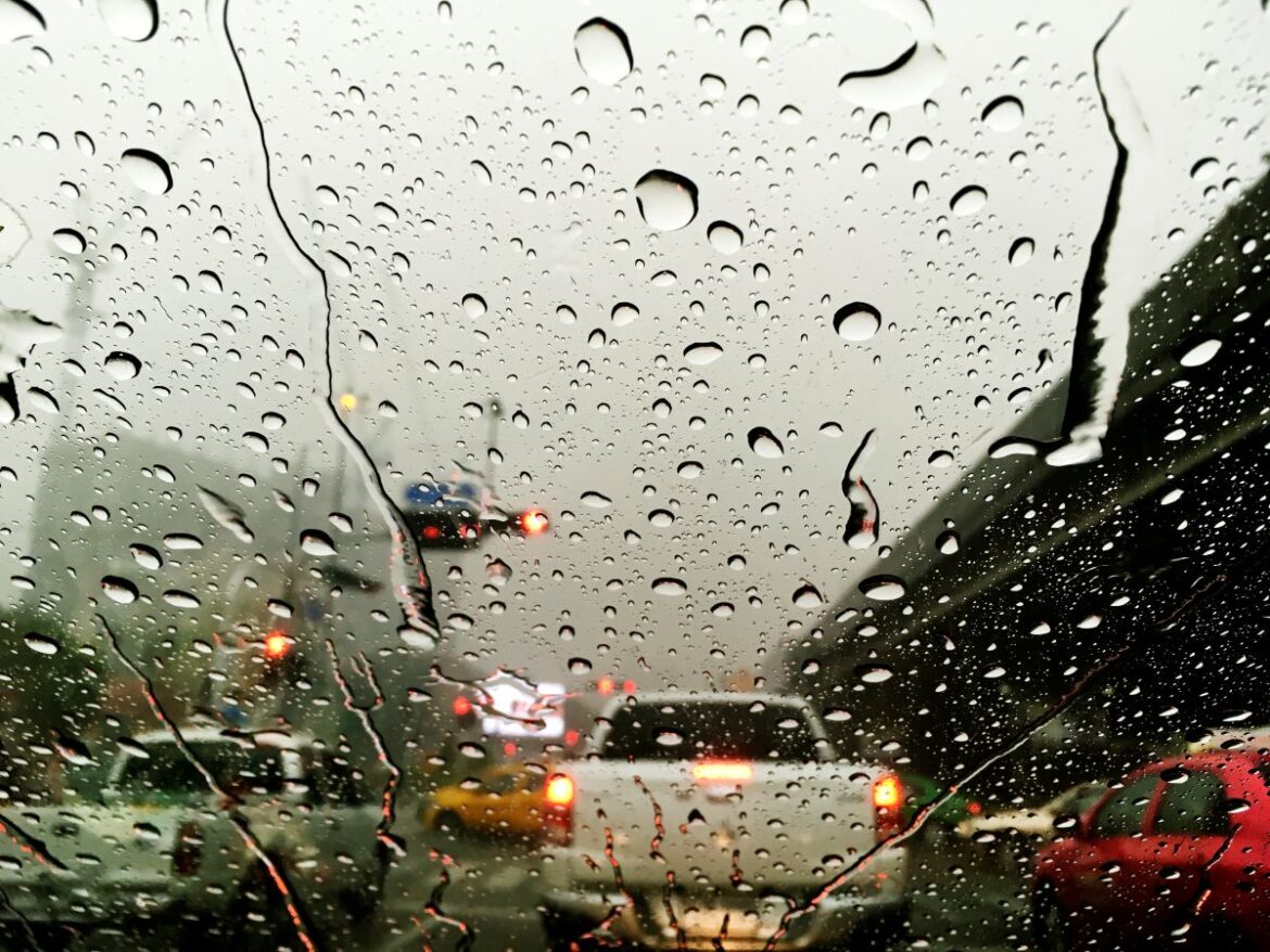 Gotas de chuva escorrendo no para-brisa de um carro, embaçando a visão de veículos em um engarrafamento sob chuva intensa. Cenário de tempo severo e mau tempo.