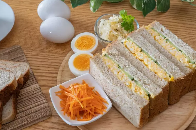 Sanduíche de Ovo Cremoso: 3 receitas fáceis e sabor imbatível para seu lanche da tarde 8 Fatias de sanduíche recheadas com ovo, cenoura ralada e abacate ao lado de ovos cozidos em uma tábua