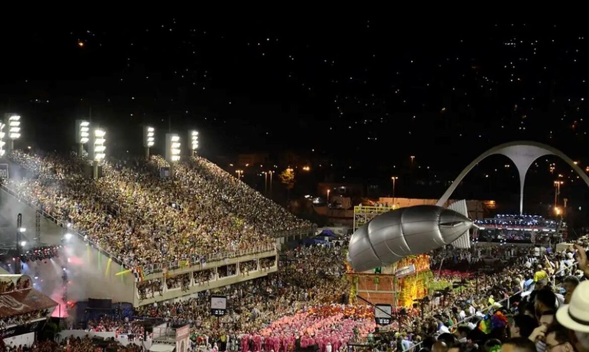 Vista panorâmica noturna do Sambódromo da Marquês de Sapucaí lotado durante o Carnaval do Rio 2026. A imagem mostra as arquibancadas iluminadas repletas de público, um carro alegórico monumental com um dirigível prateado acoplado e o icônico arco da Praça da Apoteose ao fundo sob um céu estrelado.