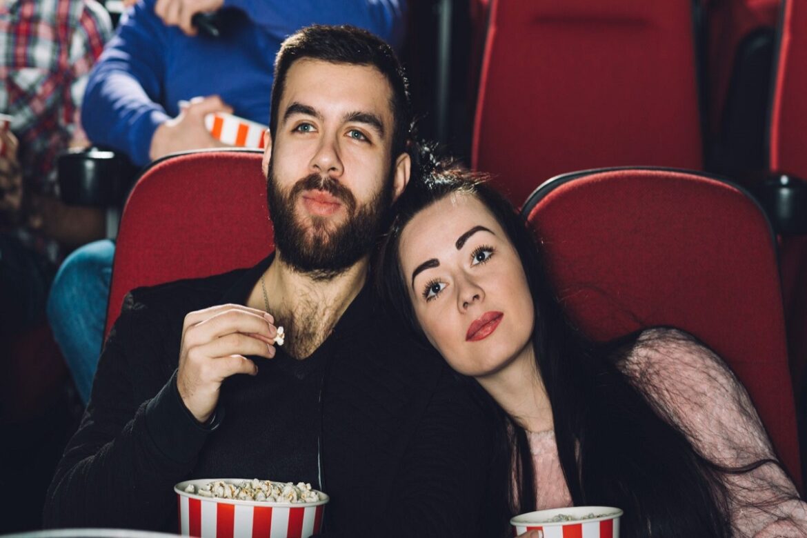 Casal sentado junto em poltronas vermelhas do cinema, segurando baldes de pipoca e assistindo filme