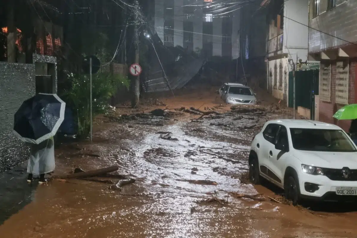 Chuva intensa causa destruição em Juiz de Fora (MG); veja imagens impressionantes 6 Uma rua urbana devastada em Juiz de Fora; um homem de costas segura um guarda-chuva observando a via coberta por lama, destroços de madeira e um carro branco parcialmente submerso sob chuva persistente.