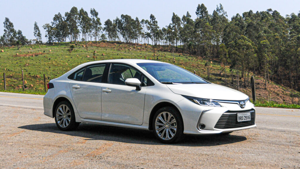 Os 5 modelos de carros usados que menos perderam valor em 2025 6 Carro sedan branco estacionado em acostamento com campo gramado e árvores ao fundo